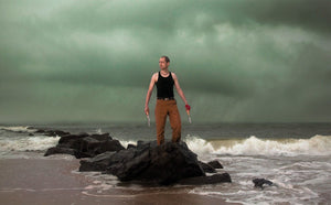 James Leonard - photo of artist standing on rock ocean breaker while storms brews in background, posed holding a carving knife in each hand