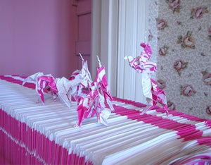 James Leonard - Detail of Hungry Dust origami  unicorns posed atop a stack of pink posters of the same material