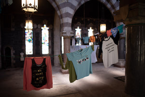 Several casual T-shirts and tank tops with custom texts hang on a clothesline strung across the interior of a stone cathedral with arched windows and hanging lanterns. Hoboken, NJ. Art by Molly Gallentine & James Leonard.