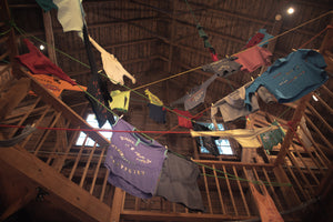The central vortex from The T-Shirt Shop at the End of Civilization. A collection of colorful graphic t-shirts hanging from colored lines within the  wooden loft of a barn, photographed from a low angle looking up. Artists James Leonard and Molly Gallentine. Sudbury, MA 2026.