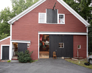 A large red barn with white trim and dark gray doors, one of which is open to show The T-Shirt Shop at the end of Civilization as its interior. Trees and a paved driveway surround the structure. Artists Molly Gallentine and James Leonard. Sudbury MA, 2026
