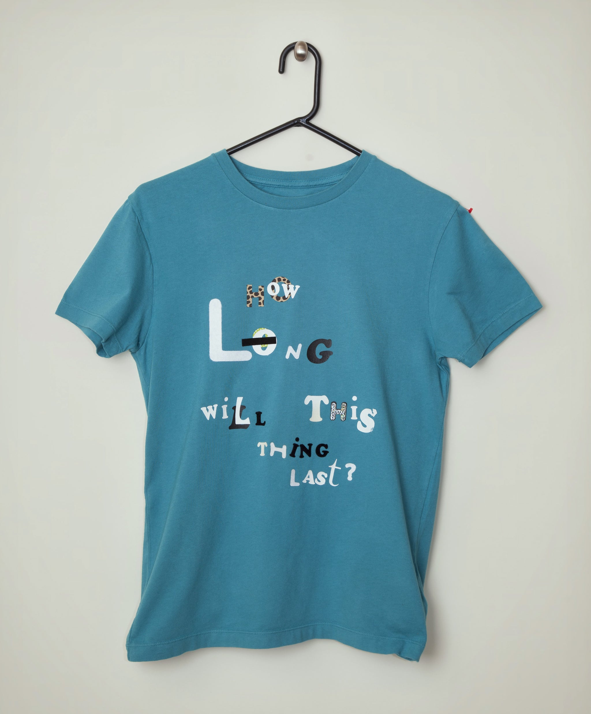 A teal blue short-sleeve t-shirt from The T-Shirt Shop at the End of Civilization hanging on a black plastic hanger against a plain. The shirt features a graphic text print on the front reading 'LONG WILL THIS THING LAST?' in stylized white and black lettering.
