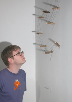 James Leonard - A man stands to examine Running Fence no. 2, a gallery installation of a miniature, handmade barbed  wire fence; it begins at his eye level, traveling up a wall