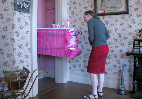 James Leonard - A woman looking at Hungry Dust, a stack of pink, flocked unicorn posters extending horizontally from a wall and held in place by two nylon bands, with origami unicorns resting on top