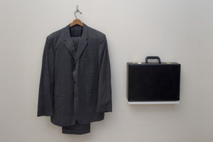 James Leonard - Costuming from Sky & Culture performance: gray suit hung on silver hook, situated to the left of a black briefcase which sits upon a shelf at sane height