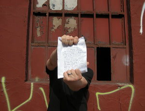 James Leonard - Photo or artist holding pages of unSuicide Note in front of his face; he stands against an exterior red wall in an urban area